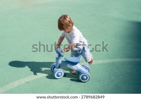 A tiny toddler boy concentrations on mounting a blue four-wheeled balance bike on an outdoor sports court. The little child in a white t-shirt and light shorts steps over the seat, casting a distinct dark shadow on the green rubber flooring next to a painted tactical marking line.