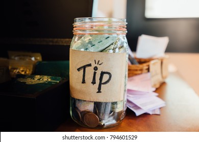 Tip box, money bank and coins in the vintage glass on wood counter in cafe