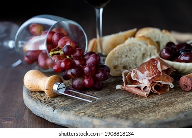 Tipped Over Wine Glass With Red Grapes Spilling Out Onto A Rustic Board Surrounded By Bread, Olives And Prosciutto.