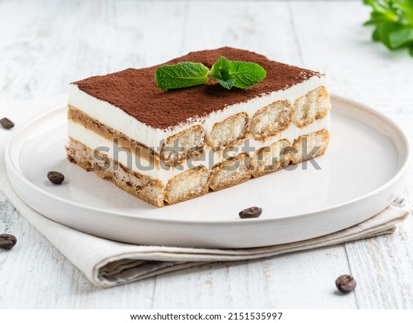 Tiramisu cake decorated with cocoa powder and
fresh green mint leaf on white ceramic plate. Close up food.
Traditional italian dessert. Coffee beans, textile napkin, white
wooden table
background.