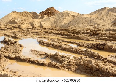 Tire Track On The Wet Soil Near The Construction Side. Construction Dirt Pile. Large Hill Of Sandy Soil With Vehicle Tire Tracks On The Surface. Blue Sky And Clouds Background.