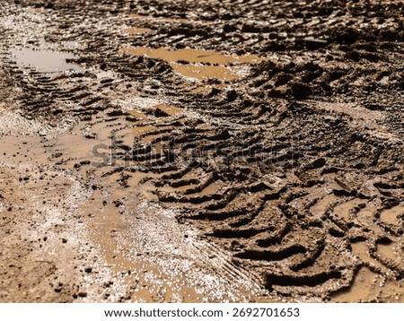 Tire tracks create intricate patterns in wet mud at a rural construction site. Puddles form in the low areas, reflecting the sky. The scene highlights the effects of rain on the terrain