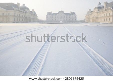 Tire tracks create linear patterns across a frozen snow-covered landscape, leading toward elegant palatial buildings in the distance.