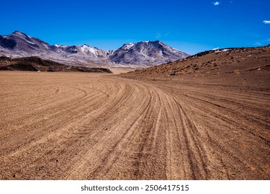 Tire tracks curve through a dry, desert landscape in Bolivia, leading towards snow-capped mountains under a vibrant blue sky. The image captures the rugged, remote beauty of this high-altitude region. - Powered by Shutterstock