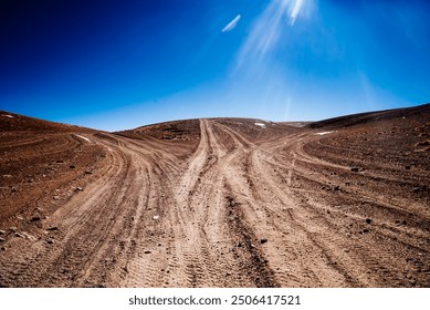 Tire tracks curve through a dry, desert landscape in Bolivia, leading towards snow-capped mountains under a vibrant blue sky. The image captures the rugged, remote beauty of this high-altitude region. - Powered by Shutterstock