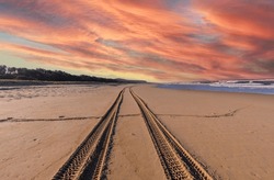 Tire Tracks On The Sandy Beach Leading Into The Horizontal Sea With Golden Sunrise Light Rey Effect On Horizon View Background.