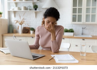 Tired 30s Hispanic woman sit in kitchen at home takes off eyeglasses, rubs nose bridge reducing discomfort after long usage of wireless computer. Blurry vision, eyes train, dry eyes need drops concept