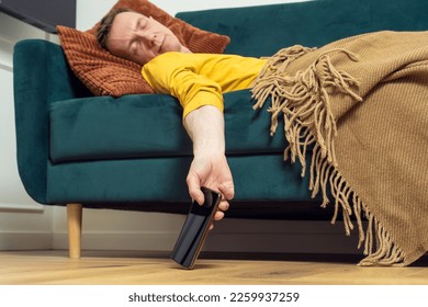 Tired adult man sleep on sofa with smartphone in hanging hand in living room. Overworking person reclining on pillow under blanket, from below view. Home relaxation, rest break, leisure. - Shutterstock ID 2259937259