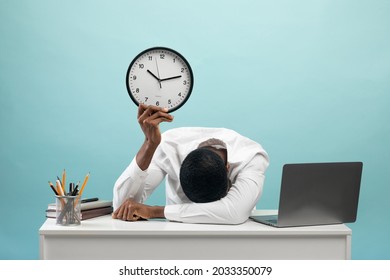 Tired african american businessman sitting at desk with pc laptop, holding clock and putting head on hand, feeling exhausted over blue studio background