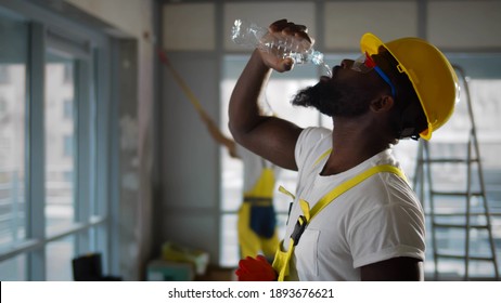 Tired African American Construction Worker Drinking Water. Side View Of Afro Builder In Safety Uniform And Hardhat Drinking Bottle Of Water Renovating Apartment Or Office