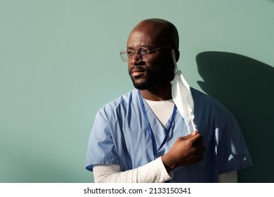 Tired African American Doctor In Blue Uniform Putting Off Protective Mask After Hard Working Day While Standing Against Wall