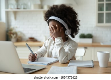 Tired African American little girl wearing headphones studying online, sitting at table in modern kitchen, exhausted child schoolgirl watching webinar, listening to lecture, homeschooling concept