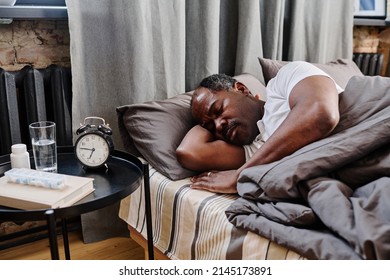 Tired African American male pensioner napping under gray blanket in bed in front of small table with alarm clock and medicaments