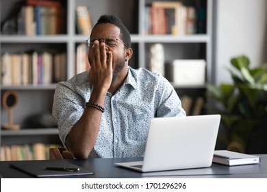 Tired african American male worker or student sit at desk sigh yawn feeling stressed or fatigue overwork in office, exhausted biracial man suffer from insomnia, need sleep or rest, exhaustion concept