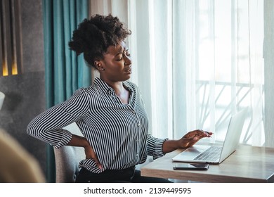 Tired african American millennial female worker sit at desk touch back suffer from lower spinal spasm, hurt unhealthy biracial woman stretch have strong backache, incorrect posture concept