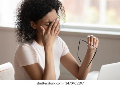 Tired african american millennnial woman dropping eyeglasses, rubbing eyes, feeling exhausted after long working day with computer, suffering from eye strain, blurred vision, dry eyes syndrome.