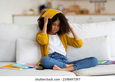 Tired African American School Girl With Bushy Hair Sitting On Couch Among Books, Notepads, Stressed Kid Pupil Doing Homework Alone, Holding Notepad On Her Head, Copy Space. Difficulties With Education