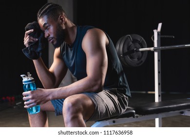 tired african american sportsman holding towel and bottle of water while sitting in gym - Powered by Shutterstock