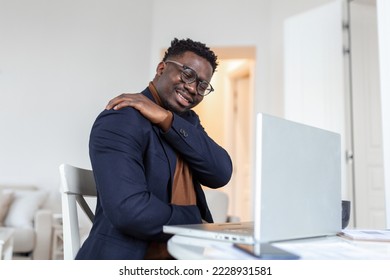 Tired African businessman working from home on his computer, feeling pain in his shoulder. man suffering from shoulder and back pain while sitting and working from home on laptop. - Powered by Shutterstock