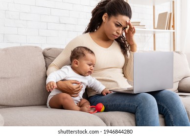 Tired African Mother With Crying Baby Using Laptop Having Headache And Trying To Work Distantly Online Sitting On Sofa At Home. Sick Exhausted Mom Browsing Internet On Computer Caring For Child