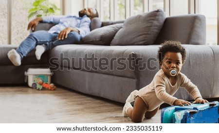Tired African-American father naps on the couch because of the necessity of raising his naughty son, enjoying toys. Father works hard. Exhausted, he returns home to take care of his son at home.
