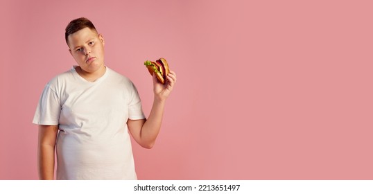 Tired Annoyed Overweight Teen Boy In White T-shirt Refuses To Eat Big Burger Isolated On Pink Background. Fast Food, Taste, Body Positive, Dieting And Facial Expressions. Copy Space For Ad