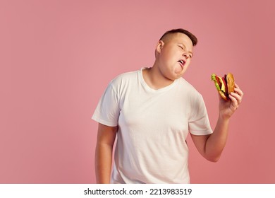 Tired Annoyed Overweight Teen Boy In White T-shirt Refuses To Eat Big Burger Isolated On Pink Background. Fast Food, Taste, Body Positive, Dieting And Facial Expressions. Copy Space For Ad