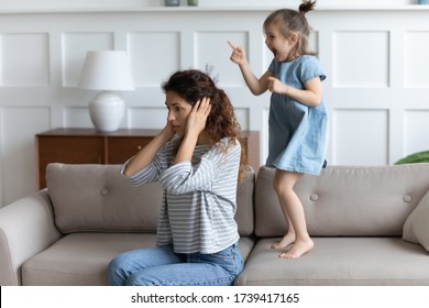 Tired Annoyed Single Mother Covering Ears, Having Problem With Naughty Little Daughter Screaming And Jumping On Couch, Child Tantrum Manipulation Concept, Preschool Noisy Demanding Attention