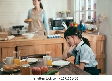Tired Asian Chinese Little Girl In Uniform Before School Fell Asleep In Early Morning Waiting For Breakfast Not Enough Sleep. Elegant Wife Mom In Apron Busy Prepare Handmade Delicious Healthy Meal