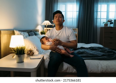 Tired Asian Father Sitting At Bedside With Eyes Closed Is Humming Lullaby While Holding And Feeding His Baby Girl Milk From Bottle In Dark Bedroom Late At Night