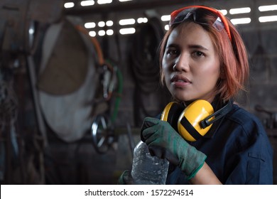 Tired Asian Female Workshop Engineer Holding Water Bottle Wearing Industrial Safety Equipment In Factory - Young Engineering Apprentice Woman Taking Break With Copy Space -Industry And Trainee Concept