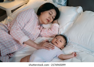 Tired Asian First Time Mom Keeping Hand On The Waking Baby’s Chest Is Falling Asleep While Putting It To Sleep On Bed In The Bedroom At Night.