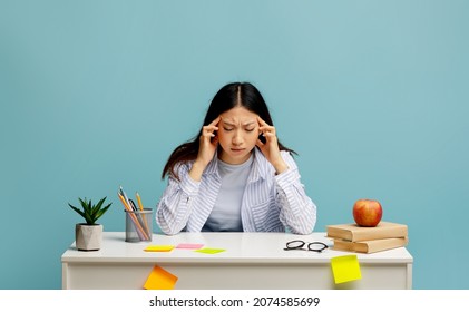 Tired Asian Lady Feeling Headache Or Stress, Massaging Her Temples, Trying To Learn And Study, Doing Homework And Thinking, Sitting At Desk Over Blue Background