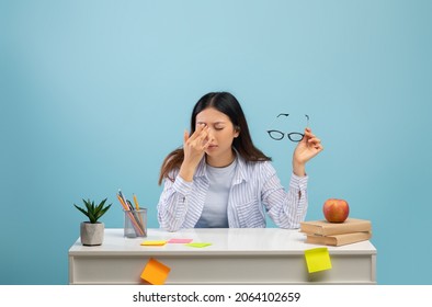 Tired Asian Lady Massaging Dry Irritated Eyes While Studying, Preparing For Exams, Sitting At Desk Over Blue Studio Background, Free Copy Space