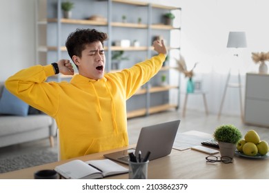 Tired Asian Male Student Yawning Sitting At Desk Using Laptop. Bored Guy Is Exhausted From Getting Ready For Test Or Writing Coursework, Feeling Sleepy, Stretching Arms. Lack Of Sleep And Tiredness