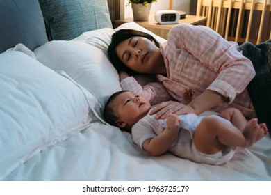 Tired Asian New Mom Wearing Pajamas Is Falling Asleep On The Bed While Comforting Her Waking Baby At Night During Bedtime In The Bedroom.