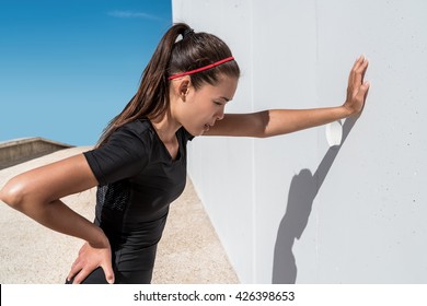 Tired athlete runner exhausted of cardio workout breathing hard after difficult exercise. Asian fitness woman running sweating of heat exhaustion leaning on wall of muscle back pain or cramps.