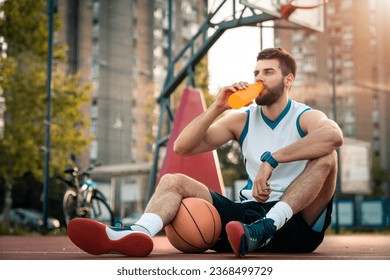 Tired basketball player drinking water from bottle while sitting at basketball court outdoors. Young man taking a break from sports activity rehydrating his body. - Powered by Shutterstock