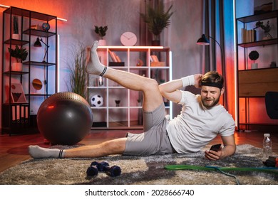 Tired Bearded Man In Sport Clothes Lying On Floor With One Leg Up. Caucasian Guy Training During Evening Time At Home. Using Smartphone For Workout.