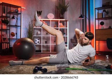 Tired Bearded Man In Sport Clothes Lying On Floor With One Leg Up. Caucasian Guy Training During Evening Time At Home. Using Smartphone For Workout.