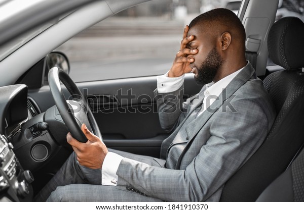 Tired black businessman having troubles at work,\
sitting in car, touching his face, side view, copy space. Exhausted\
african american entrepreneur having headache, suffering from\
stress at office