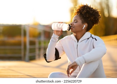 Tired Black Female Jogger Drinking Water While Relaxing After Running Outdoors, Young African American Woman Listening Music In Wireless Headphones And Enjoying Refreshing Drink, Copy Space - Powered by Shutterstock