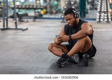 Tired black guy bodybuilder sitting on floor at gym, listening to music, using mobile phone and wireless headset, copy space. African american young man with headphones holding smartphone - Powered by Shutterstock