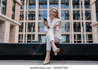 Tired blonde caucasian businesswoman sitting against blurry building thinking, looking aside. Italian pretty student girl waiting for exam results outdoors. Female entrepreneur having coffee break.