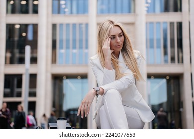 Tired blonde caucasian businesswoman sitting against blurry building thinking, looking aside. Italian pretty student girl waiting for exam results outdoors. Female entrepreneur frustrated by failure.