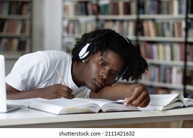 Tired Bored Lazy Black Student Listening To Audio Lesson, Webinar, Writing Notes, Reading Open Book In Library With Head Rested On Table. Overworked Exhausted College Guy Working On Study Project