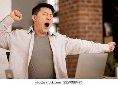 Tired bored middle aged asian man yawning while sitting at workplace with computer, wants to sleep at home interior. Work and business with modern technology, fatigue and overwork, deadline
