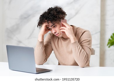 Tired Bored Sad Guy, Young Handsome Latino Ethnic Man Is Talking On Cell Mobile Phone, Having Boring Negative Conversation Talk On His Smartphone, Looking At Laptop Computer, Holding Head With Hand