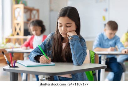 Tired and bored schoolgirl sitting at desk in classroom at school, writing in her notebook and thinking, resting head on hand - Powered by Shutterstock