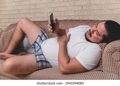 A tired and bored slacker in boxer shorts is addicted on the phone during a lazy day lying on the couch.